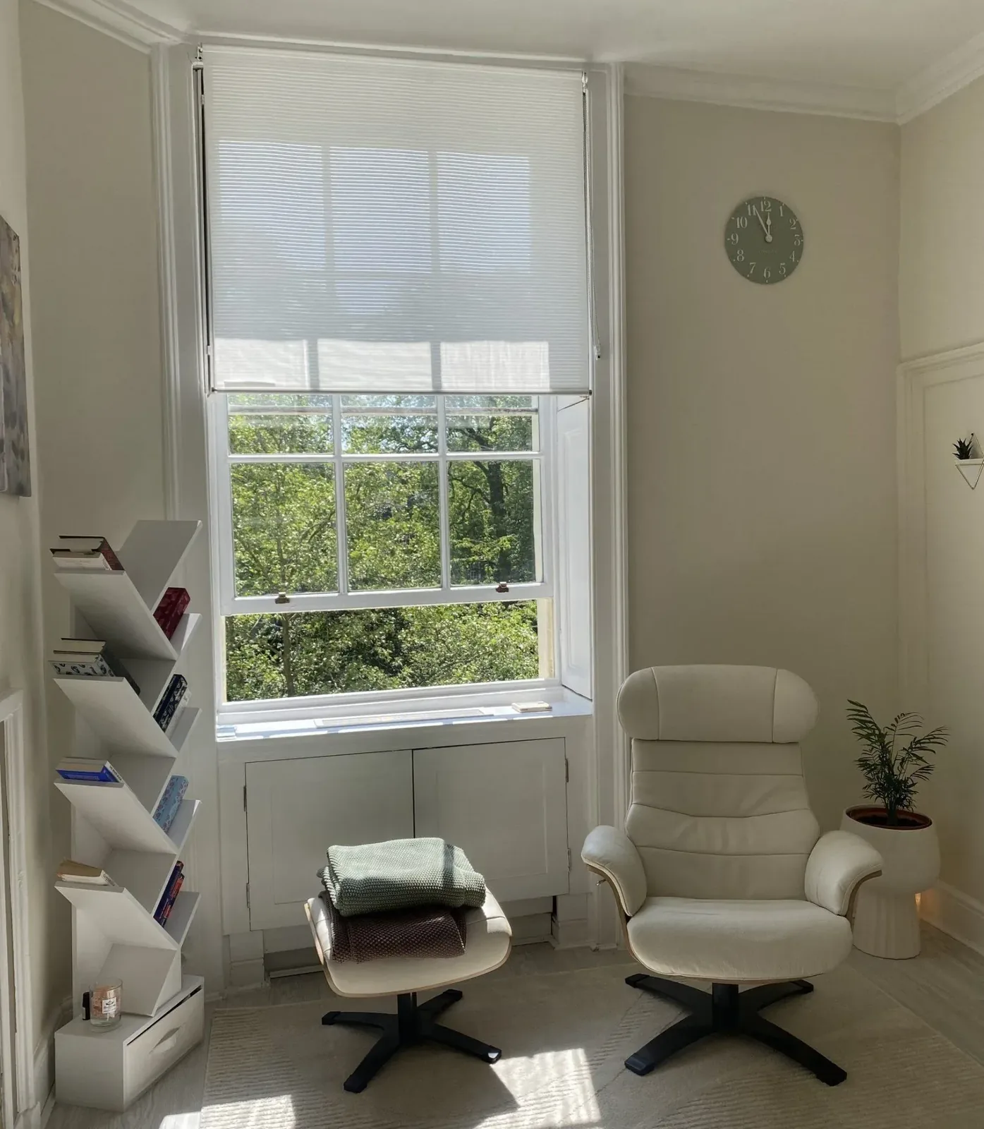 A cream armchair and footstool by a tall sash window, with a plant and a shelf of books